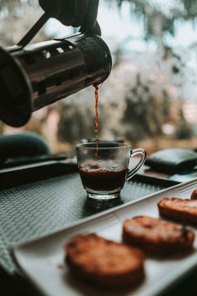 Aromatic coffee being poured into a glass cup beside a plate of biscotti on a tray.